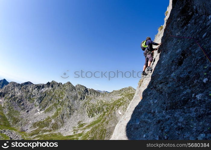 Caucasian male climber climbing a steep wall. In background a summer alpine landscape. Clear sky, day light. West italian Alps, Europe.