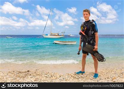 Caucasian male diver stands on beach of Bonaire with sea and boats