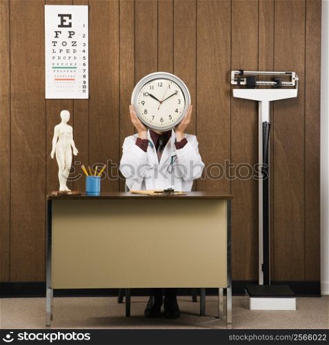 Caucasian male doctor sitting at desk holding clock over face.