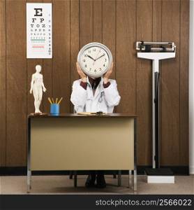 Caucasian male doctor sitting at desk holding clock over face.