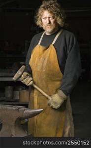 Caucasian male metalsmith holding hammer with stern expression.