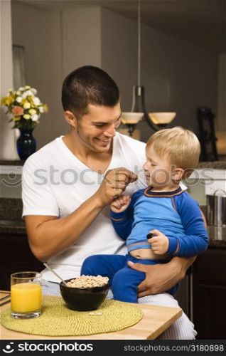 Caucasian man feeding toddler son on lap in kitchen.