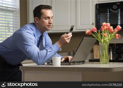 Caucasian man in suit using laptop computer and cellphone in kitchen.