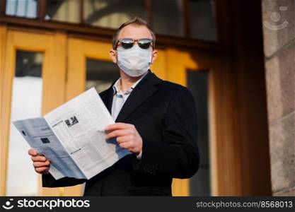 Caucasian man in sunglasses, medical mask and formal black suit, stands near building outdoor, holds newspaper in hands, poses at street, walks in quarantined city, finds out news from press