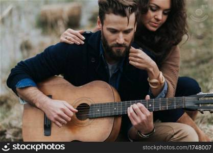 caucasian man is playing guitar with woman by the lake. Young couple is hugging on autumn day outdoors. A bearded man and curly woman in love. Valentine’s Day. Concept of love and family. caucasian man is playing guitar with woman by the lake. Young couple is hugging on autumn day outdoors. A bearded man and curly woman in love. Valentine’s Day. Concept of love and family.