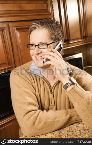 Caucasian man smiling and talking on cellphone while leaning on kitchen counter.