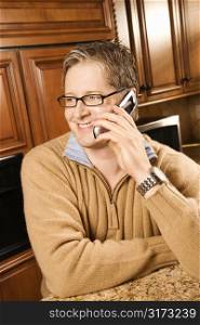 Caucasian man smiling and talking on cellphone while leaning on kitchen counter.