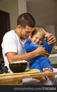 Caucasian man tickling toddler son in kitchen.