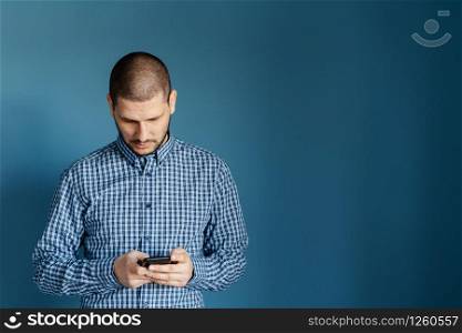 Caucasian man wearing shirt standing in front of the blue background wall using smart phone mobile to send messages sms texting or browsing internet front view