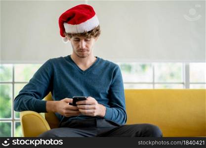 Caucasian man with Christmas hat use mobile phone and sit on sofa in his house alone during pandemic of Covid-19 to stay at home with new normal lifestyle.