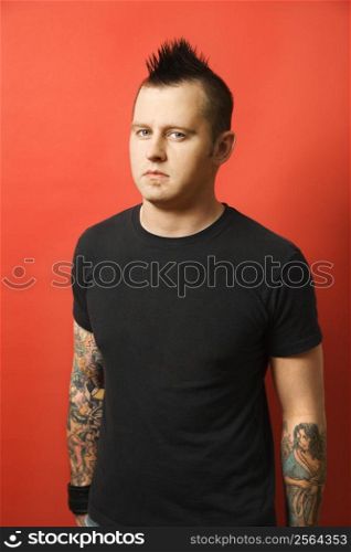 Caucasian man with mohawk and tattoos standing against orange background.