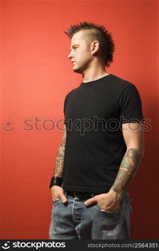 Caucasian man with mohawk and tattoos standing with hands in pockets against orange background.