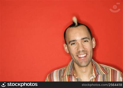 Caucasian man with mohawk smiling against red background.