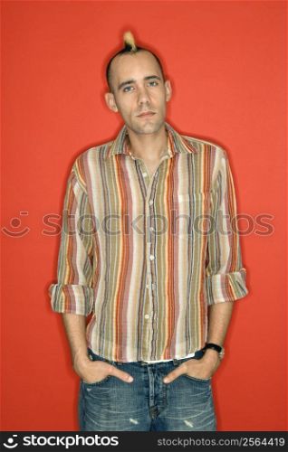 Caucasian man with mohawk standing against red background.
