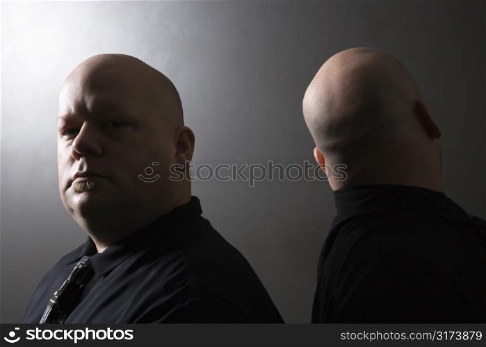 Caucasian mid adult bald identical twin men standing back to back and looking at viewer.