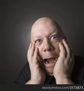 Caucasian mid adult bald man with hands to face making scared facial expression.