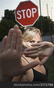 Caucasian mid-adult blonde woman holding hands up to camera in front of stop sign.
