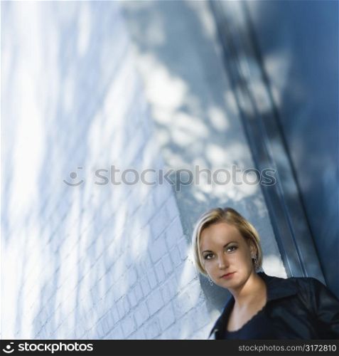 Caucasian mid-adult blonde woman standing against sun dappled building looking at viewer.