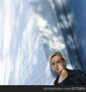 Caucasian mid-adult blonde woman standing against sun dappled building looking at viewer.