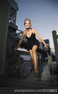 Caucasian mid-adult blonde woman wearing little black dress walking on dock along bridge.