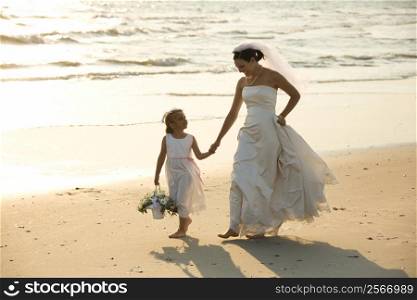 Caucasian mid-adult bride and flower girl holding hands walking barefoot on beach.