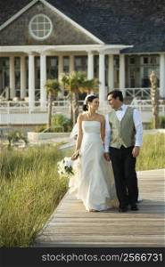 Caucasian mid-adult bride and groom walking down wooden beach walkway holding hands.