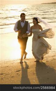 Caucasian mid-adult bride and mid-adult groom holding hands running barefoot on beach.