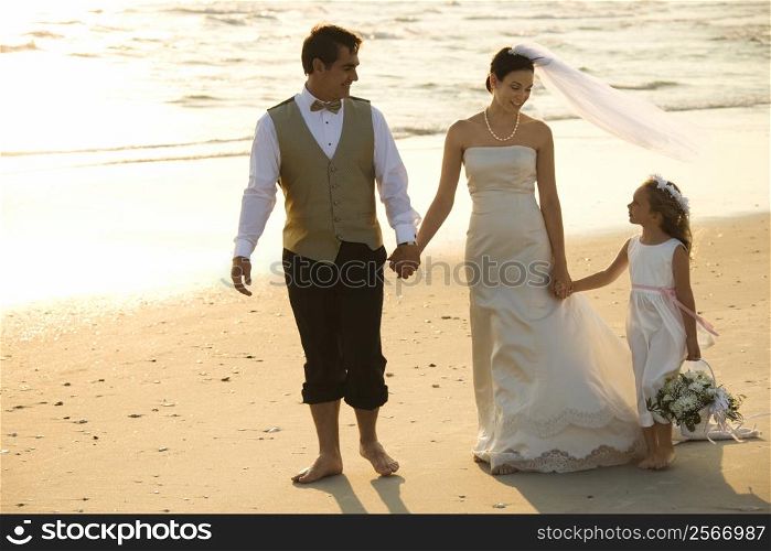 Caucasian mid-adult bride, mid-adult groom and flower girl holding hands walking barefoot on beach.