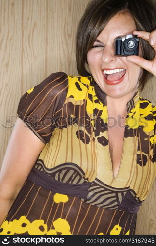 Caucasian mid adult brunette woman looking through miniature toy camera and making facial expression.