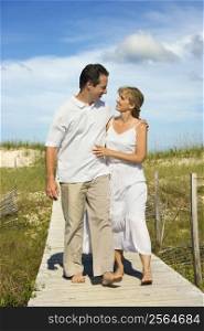 Caucasian mid-adult couple holding each other walking down beach access path.