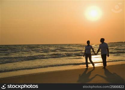 Caucasian mid-adult couple holding hands and walking down beach at sunset.