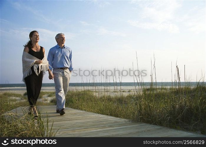 Caucasian mid-adult couple holding hands and walking down walkway at beach.