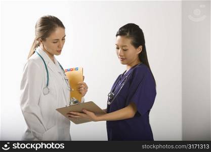 Caucasian mid-adult female doctor and Asian Chinese mid-adult female physician&acute;s assistant discussing paperwork.