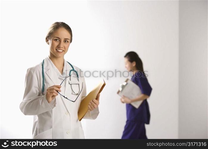 Caucasian mid-adult female doctor smiling and looking at viewer with Asian Chinese mid-adult female physician&acute;s assistant walking by in background.
