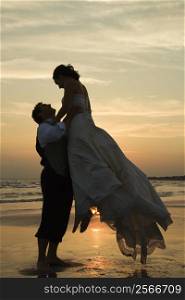 Caucasian mid-adult groom lifting up bride on beach at sunset.
