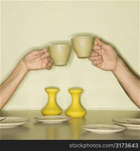 Caucasian mid-adult male and female hands toasting with coffee cups across retro kitchen table setting.