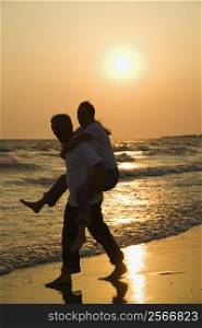 Caucasian mid-adult male carrying female piggyback on beach at sunset.