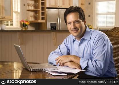 Caucasian mid-adult man paying bills on laptop computer looking at viewer and smiling.