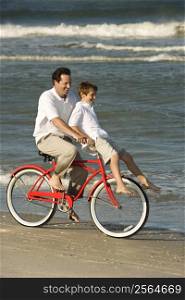 Caucasian mid-adult man riding bicycle on beach with pre-teen boy on handlebars.