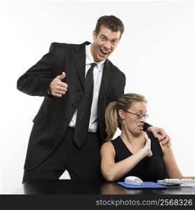Caucasian mid-adult man sexually harassing woman sitting at computer and giving thumbs up.