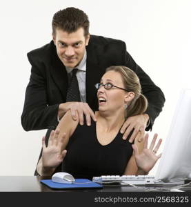 Caucasian mid-adult man sexually harassing woman sitting at computer and looking at viewer.