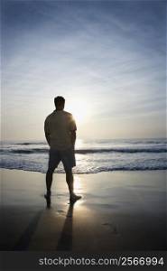 Caucasian mid-adult man standing alone on beach looking at ocean at sunrise.