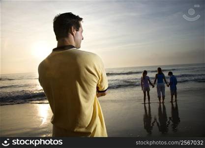 Caucasian mid-adult man standing and watching mid-adult woman with children on beach.