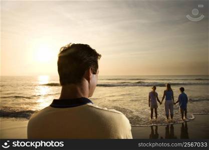 Caucasian mid-adult man standing and watching mid-adult woman with children on beach.