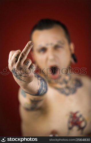 Caucasian mid-adult man with tattoos and piercings holding up middle finger.