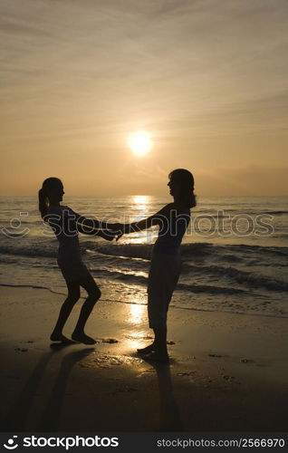 Caucasian mid-adult mother and teenage daughter holding hands spinngin on beach at sunset.