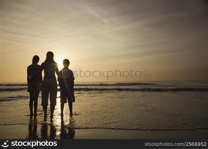 Caucasian mid-adult mother and teenage kids standing silhouetted on beach at sunset.