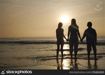 Caucasian mid-adult mother and tenage kids standing silhouetted on beach at sunset.