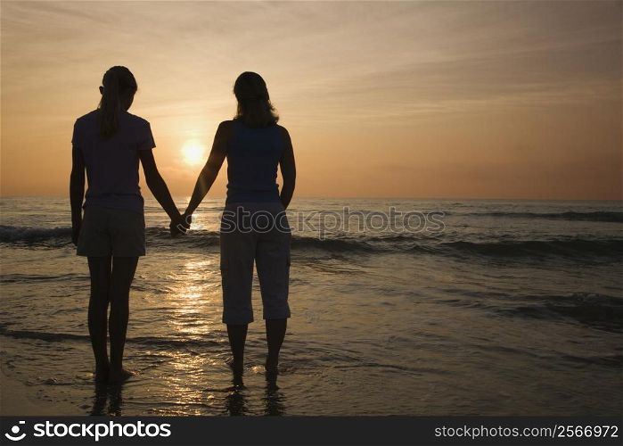 Caucasian mid-adult mother teenage and daughter standing on beach at sunset holding hands.