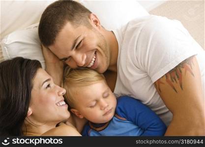 Caucasian mid adult parents cuddling with toddler son sleeping in bed.